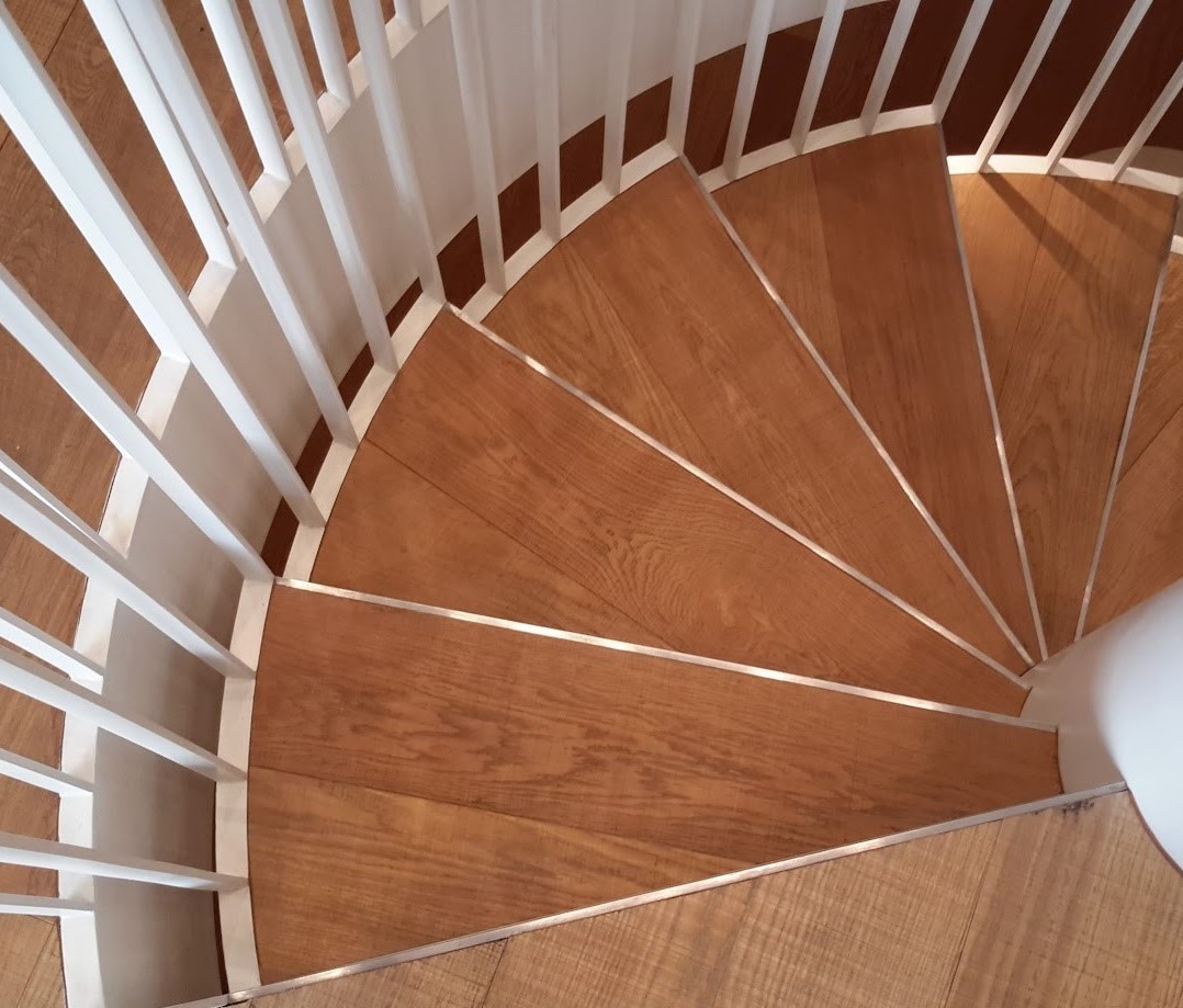 A metal spiral staircase with stair nosing and white balustrade with wooden treads, seen from above.