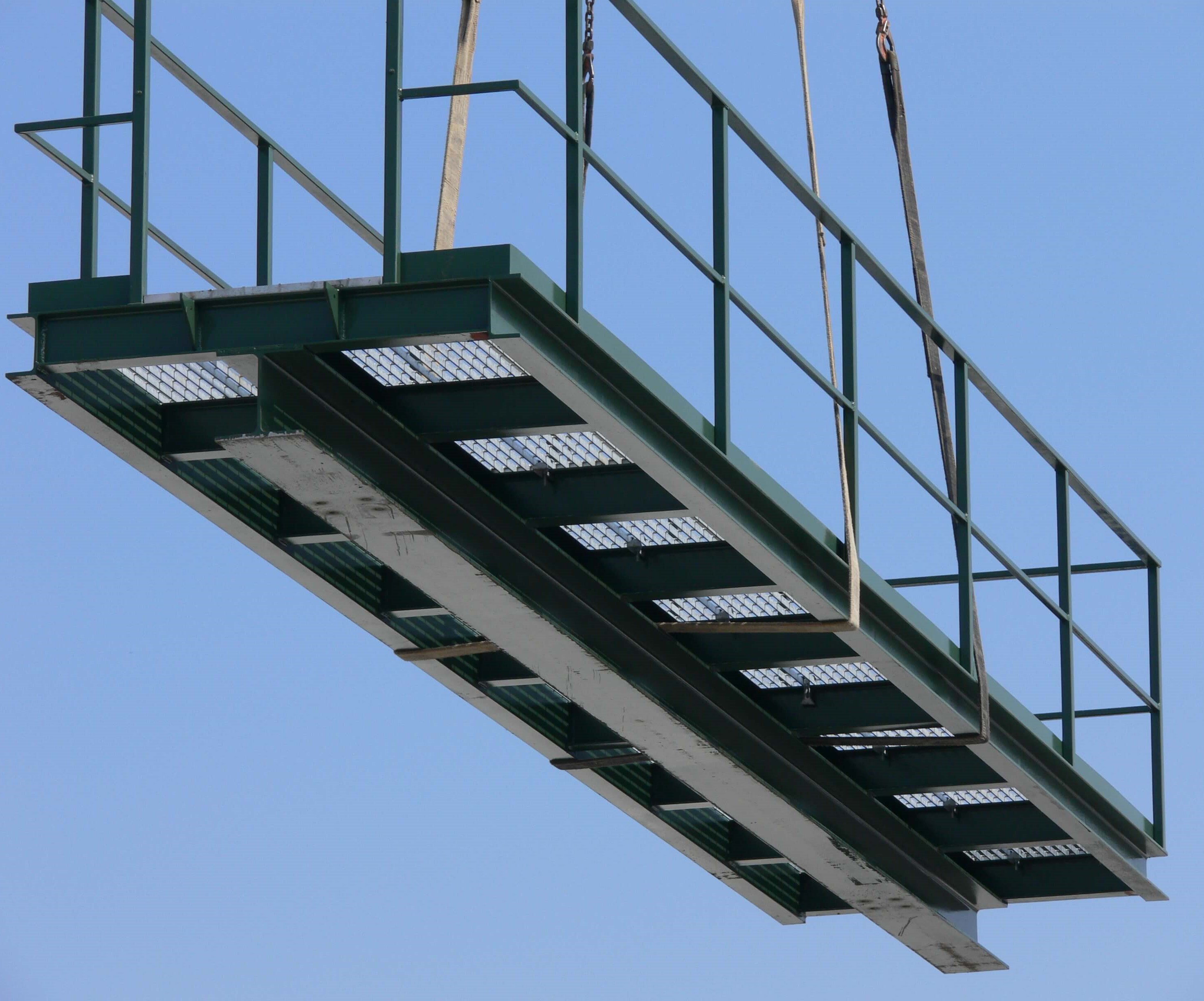 Green metal walkway with handrails suspended in the air with a crane